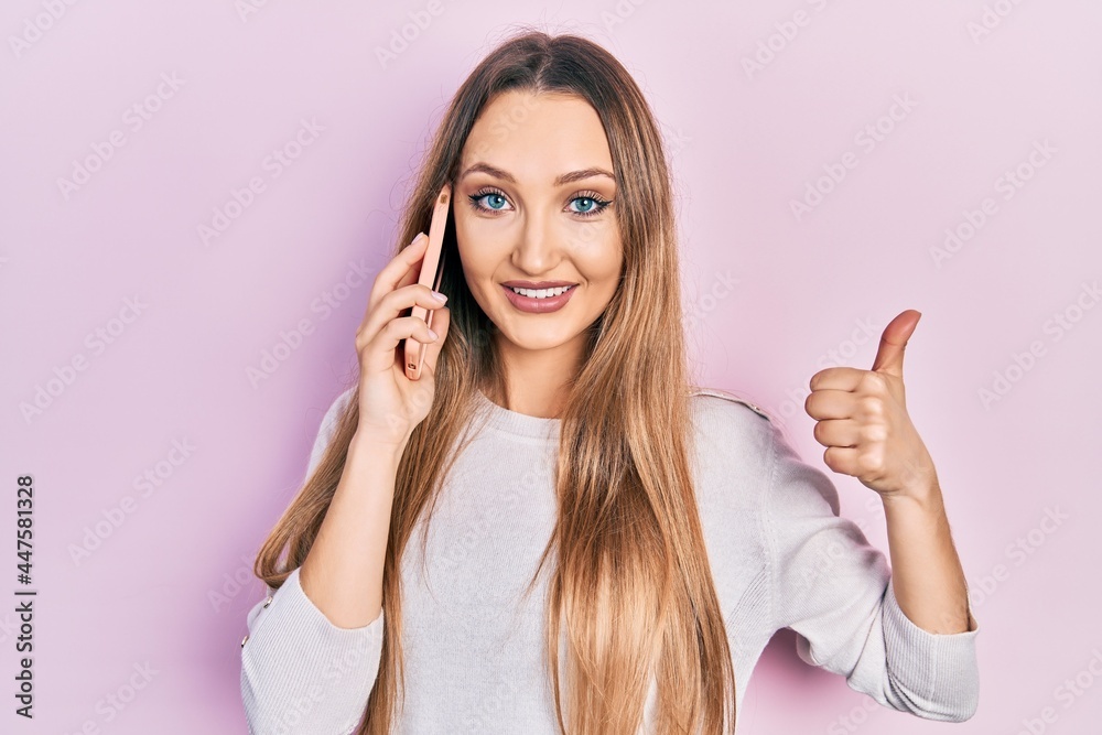 Young blonde girl having conversation talking on the smartphone smiling happy and positive, thumb up doing excellent and approval sign Young blonde girl having conversation talking on the smartphone smiling happy and positive, thumb up doing excellent and approval sign