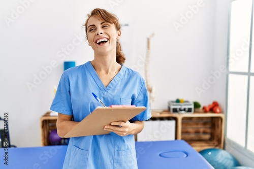 Young caucasian physio therapist girl smiling happy writing on clipboard at the clinic.