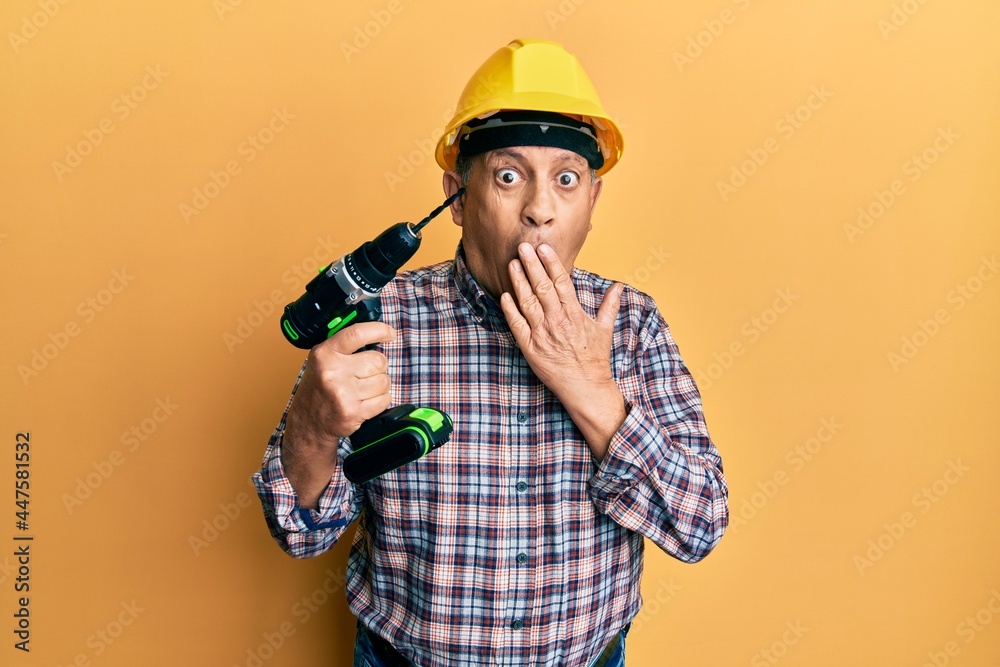 Handsome senior man with grey hair holding electric screwdriver wearing hardhat covering mouth with hand, shocked and afraid for mistake. surprised expression Handsome senior man with grey hair holding electric screwdriver wearing hardhat covering mouth with hand, shocked and afraid for mistake. surprised expression
