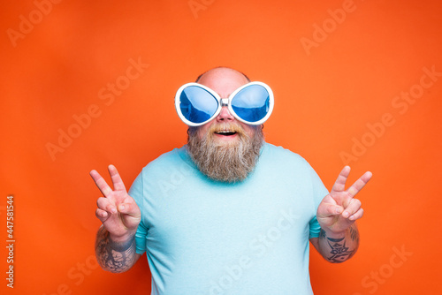 Fat happy man with beard, tattoos and sunglasses makes the sign of victory with his hands