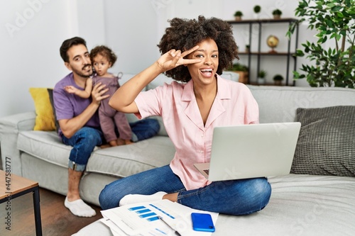 Mother of interracial family working using computer laptop at home doing peace symbol with fingers over face, smiling cheerful showing victory