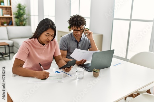 Young latin couple working using laptop and talking on the smartphone sitting on the table at home.