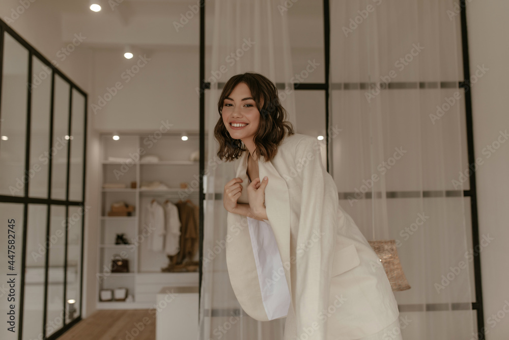 Curly short-haired brunette woman in beige trendy suit smiles widely. Young girl in jacket poses in good mood in dressing room. Curly short-haired brunette woman in beige trendy suit smiles widely. Young girl in jacket poses in good mood in dressing room.