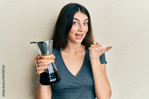 Young caucasian woman holding coffee maker with filter pointing thumb up to the side smiling happy with open mouth