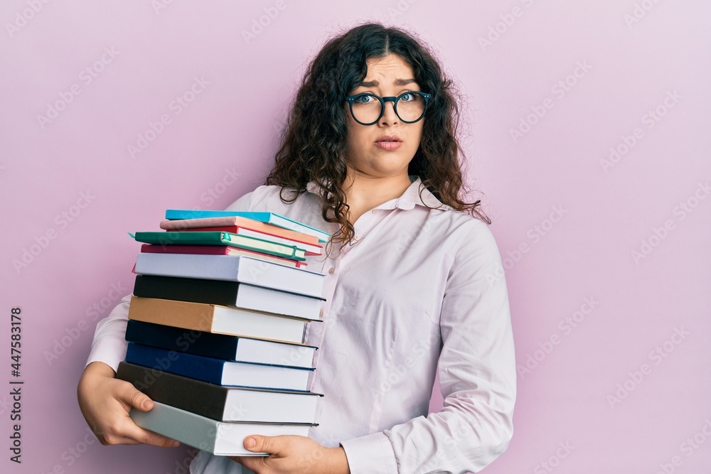 Young brunette woman with curly hair holding a pile of books clueless and confused expression. doubt concept. Young brunette woman with curly hair holding a pile of books clueless and confused expression. doubt concept.