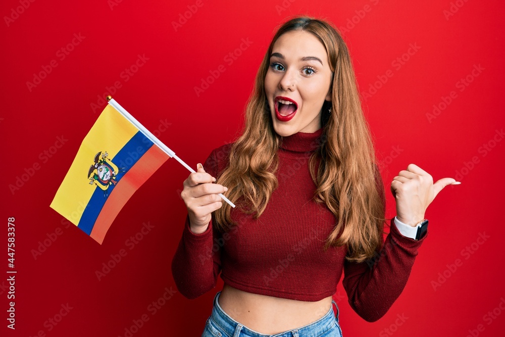 Young blonde woman holding ecuador flag pointing thumb up to the side smiling happy with open mouth