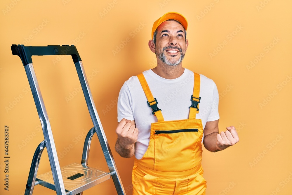 Handsome middle age man with grey hair holding ladder very happy and excited doing winner gesture with arms raised, smiling and screaming for success. celebration concept. Handsome middle age man with grey hair holding ladder very happy and excited doing winner gesture with arms raised, smiling and screaming for success. celebration concept.