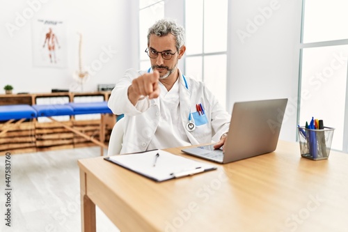 Middle age hispanic therapist man working at pain recovery clinic with laptop pointing with finger to the camera and to you, confident gesture looking serious