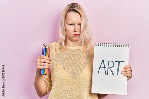 Beautiful caucasian blonde woman holding art notebook and color pencils skeptic and nervous  frowning upset because of problem. negative person.