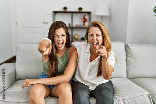 Mother and daughter together sitting on the sofa at home pointing displeased and frustrated to the camera, angry and furious with you