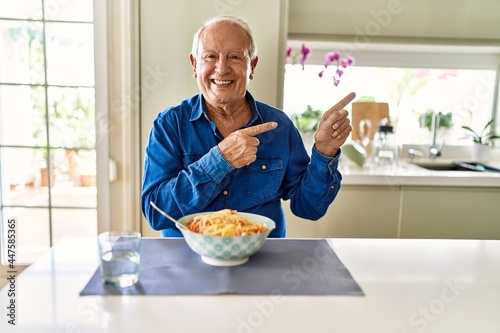Senior man with grey hair eating pasta spaghetti at home smiling and looking at the camera pointing with two hands and fingers to the side.