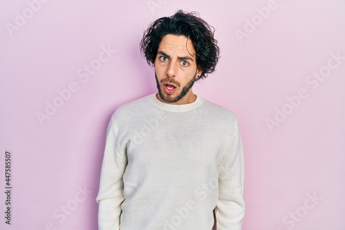 Handsome hispanic man wearing casual white sweater in shock face, looking skeptical and sarcastic, surprised with open mouth