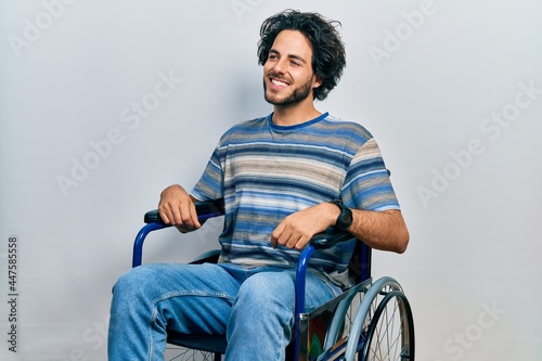 Handsome hispanic man sitting on wheelchair looking away to side with smile on face, natural expression. laughing confident.