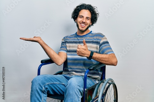 Handsome hispanic man sitting on wheelchair showing palm hand and doing ok gesture with thumbs up, smiling happy and cheerful