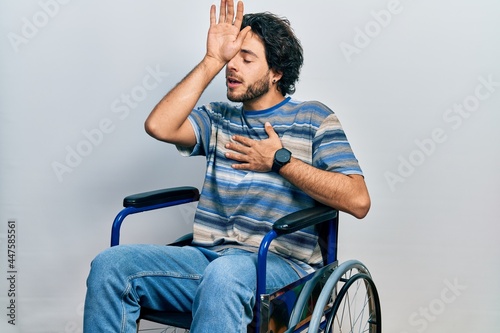 Handsome hispanic man sitting on wheelchair touching forehead for illness and fever, flu and cold, virus sick