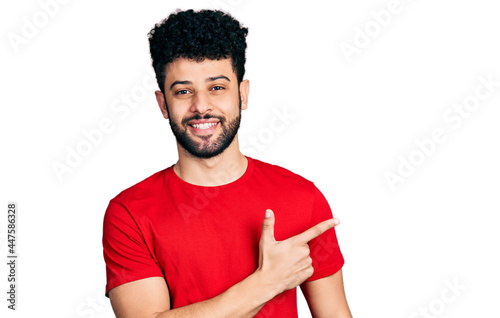 Young arab man with beard wearing casual red t shirt cheerful with a smile of face pointing with hand and finger up to the side with happy and natural expression on face