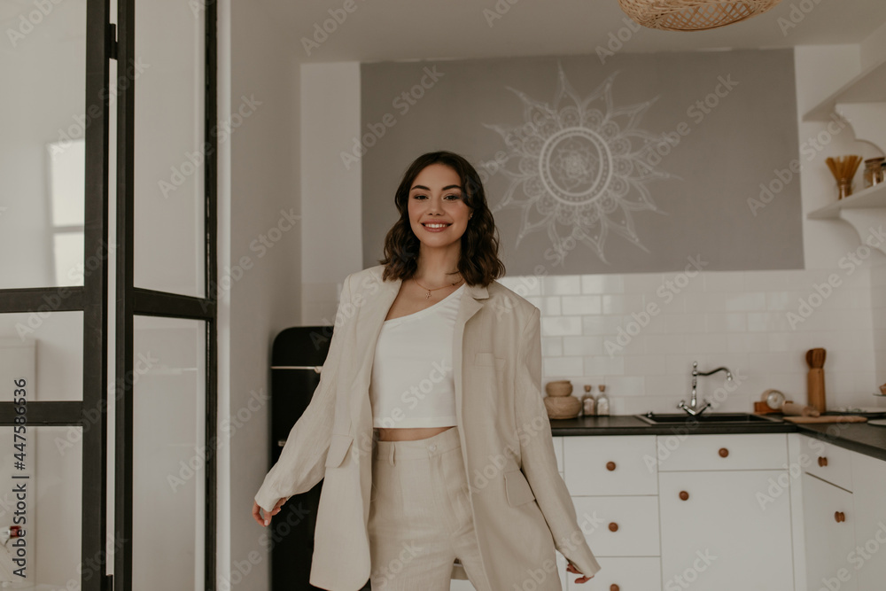 Short-haired brunette tanned woman in trendy suit smiles sincerely at kitchen. Young lady in jacket and linen pants moves at home. Short-haired brunette tanned woman in trendy suit smiles sincerely at kitchen. Young lady in jacket and linen pants moves at home.