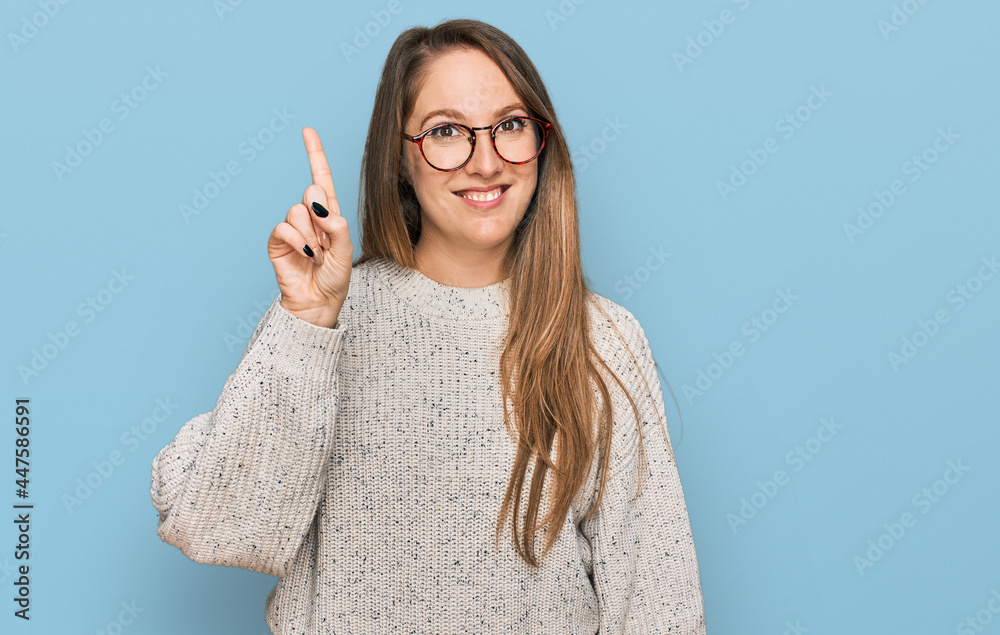 Young blonde woman wearing casual sweater and glasses showing and pointing up with finger number one while smiling confident and happy. Young blonde woman wearing casual sweater and glasses showing and pointing up with finger number one while smiling confident and happy.