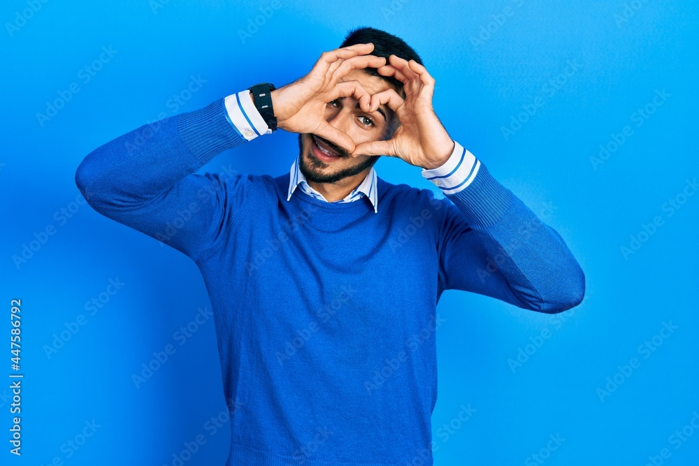 Young hispanic man with beard wearing casual blue sweater doing heart shape with hand and fingers smiling looking through sign Young hispanic man with beard wearing casual blue sweater doing heart shape with hand and fingers smiling looking through sign