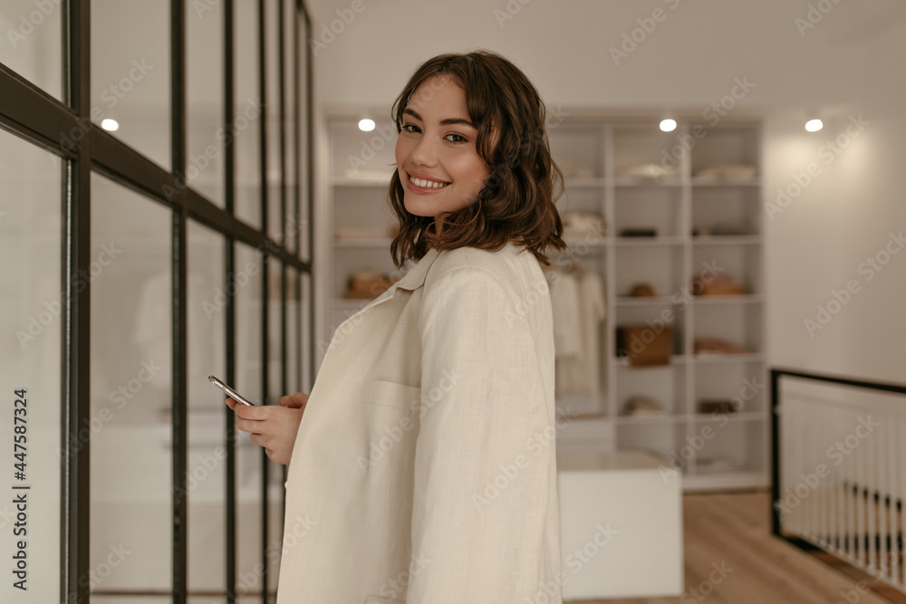 Charming brunette curly woman in trendy beige suit holds phone. Elegant young lady in linen jacket smiles sincerely and poses in dressing room.
