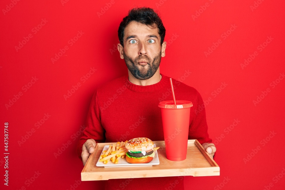 Handsome man with beard eating a tasty classic burger with fries and soda making fish face with mouth and squinting eyes, crazy and comical. Handsome man with beard eating a tasty classic burger with fries and soda making fish face with mouth and squinting eyes, crazy and comical.