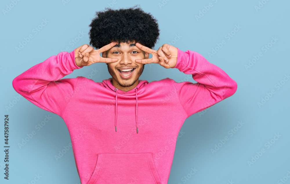 Young african american man with afro hair wearing casual pink sweatshirt doing peace symbol with fingers over face, smiling cheerful showing victory Young african american man with afro hair wearing casual pink sweatshirt doing peace symbol with fingers over face, smiling cheerful showing victory