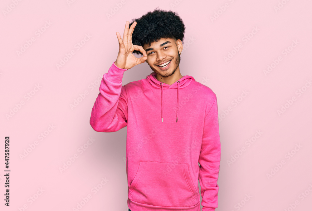 Young african american man with afro hair wearing casual pink sweatshirt smiling positive doing ok sign with hand and fingers. successful expression. Young african american man with afro hair wearing casual pink sweatshirt smiling positive doing ok sign with hand and fingers. successful expression.