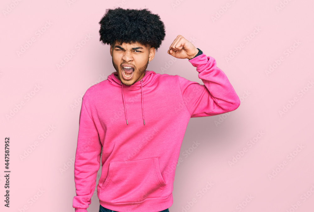 Young african american man with afro hair wearing casual pink sweatshirt angry and mad raising fist frustrated and furious while shouting with anger. rage and aggressive concept. Young african american man with afro hair wearing casual pink sweatshirt angry and mad raising fist frustrated and furious while shouting with anger. rage and aggressive concept.