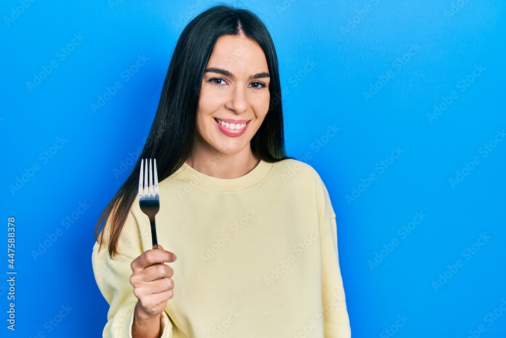 Young brunette woman holding one silver fork looking positive and happy standing and smiling with a confident smile showing teeth Young brunette woman holding one silver fork looking positive and happy standing and smiling with a confident smile showing teeth