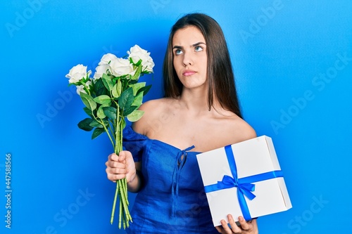 Young brunette teenager holding anniversary present and bouquet of flowers smiling looking to the side and staring away thinking.