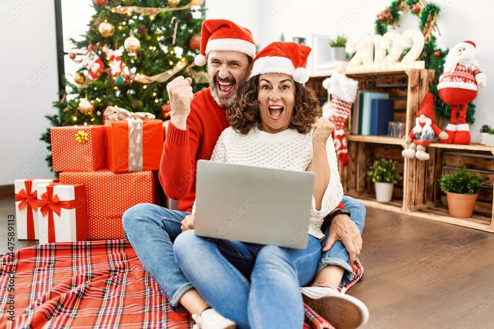 Hispanic middle age woman and mature man using laptop sitting by christmas tree screaming proud, celebrating victory and success very excited with raised arms Hispanic middle age woman and mature man using laptop sitting by christmas tree screaming proud, celebrating victory and success very excited with raised arms