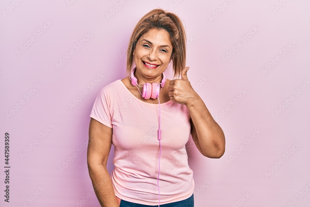 Middle age hispanic woman wearing gym clothes and using headphones smiling happy and positive, thumb up doing excellent and approval sign Middle age hispanic woman wearing gym clothes and using headphones smiling happy and positive, thumb up doing excellent and approval sign