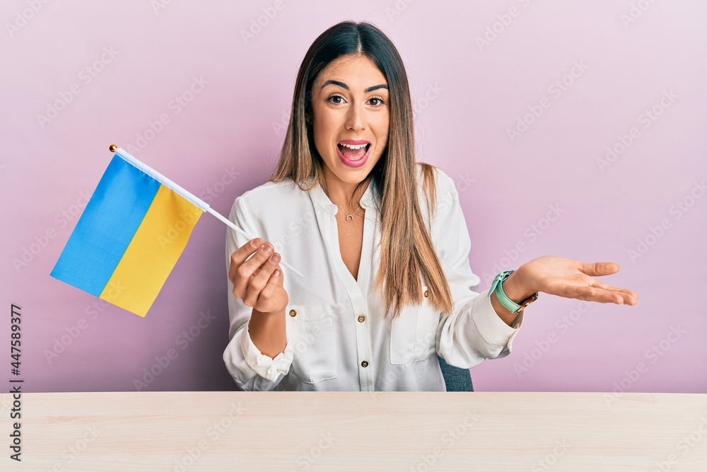 Young hispanic woman holding ukraine flag sitting on the table celebrating achievement with happy smile and winner expression with raised hand Young hispanic woman holding ukraine flag sitting on the table celebrating achievement with happy smile and winner expression with raised hand