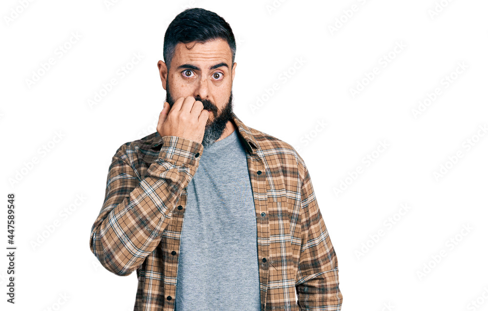 Hispanic man with beard wearing casual shirt looking stressed and nervous with hands on mouth biting nails. anxiety problem. Hispanic man with beard wearing casual shirt looking stressed and nervous with hands on mouth biting nails. anxiety problem.