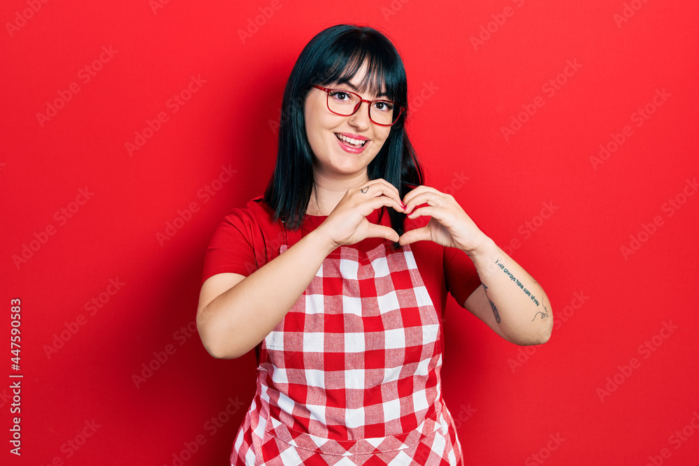 Young hispanic woman wearing cook apron and glasses smiling in love doing heart symbol shape with hands. romantic concept. Young hispanic woman wearing cook apron and glasses smiling in love doing heart symbol shape with hands. romantic concept.