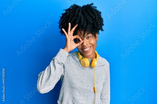African american woman with afro hair wearing sweatshirt and using headphones smiling happy doing ok sign with hand on eye looking through fingers