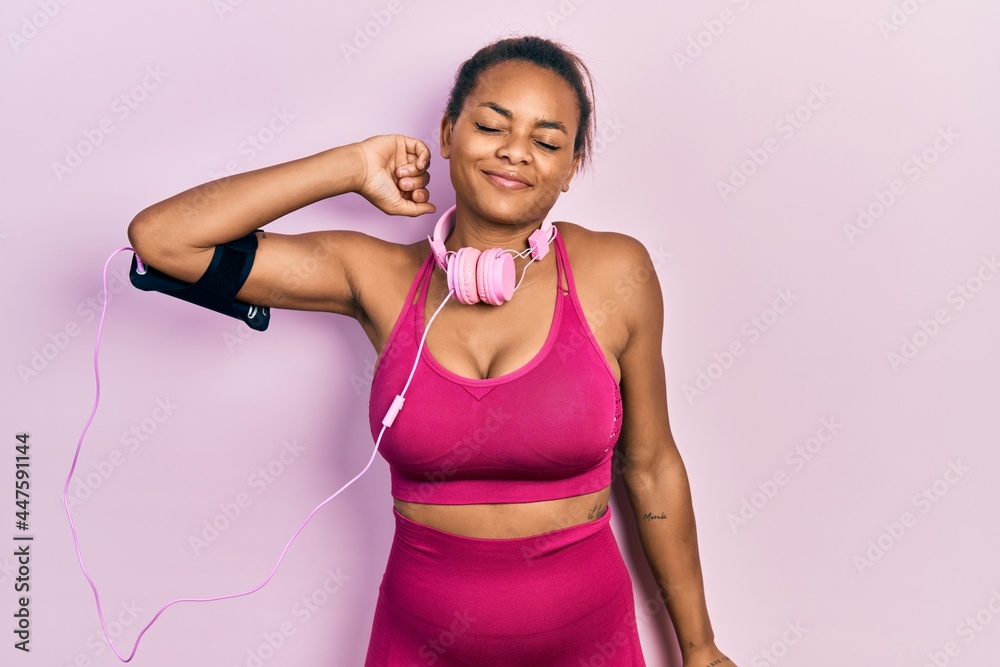 Young african american girl wearing gym clothes and using headphones stretching back, tired and relaxed, sleepy and yawning for early morning