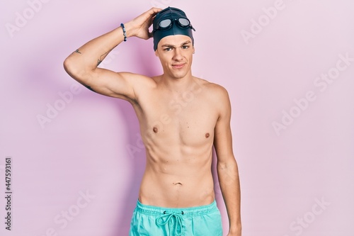 Young hispanic man wearing swimwear and swimmer glasses confuse and wonder about question. uncertain with doubt, thinking with hand on head. pensive concept. © Krakenimages.com