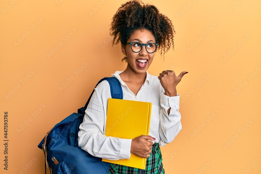 Beautiful african american woman with afro hair wearing school bag and holding books pointing thumb up to the side smiling happy with open mouth