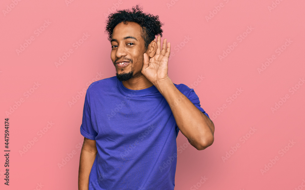 Young african american man with beard wearing casual purple t shirt smiling with hand over ear listening an hearing to rumor or gossip. deafness concept. Young african american man with beard wearing casual purple t shirt smiling with hand over ear listening an hearing to rumor or gossip. deafness concept.