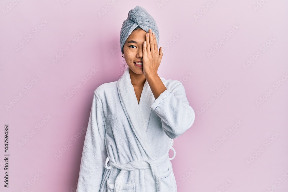 Young chinese woman wearing shower towel cap and bathrobe covering one eye with hand, confident smile on face and surprise emotion. Young chinese woman wearing shower towel cap and bathrobe covering one eye with hand, confident smile on face and surprise emotion.