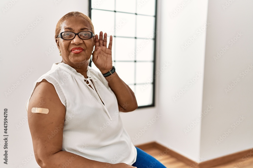 Mature hispanic woman getting vaccine showing arm with band aid smiling with hand over ear listening an hearing to rumor or gossip. deafness concept.