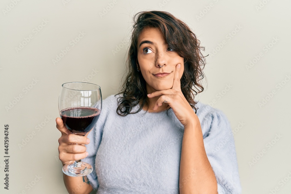 Young hispanic woman drinking a glass of red wine serious face thinking about question with hand on chin, thoughtful about confusing idea Young hispanic woman drinking a glass of red wine serious face thinking about question with hand on chin, thoughtful about confusing idea