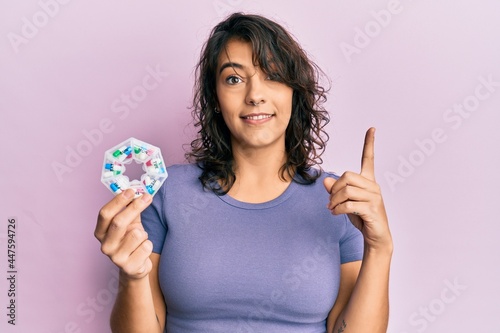 Young hispanic woman holding pill organizer smiling with an idea or question pointing finger with happy face, number one photo