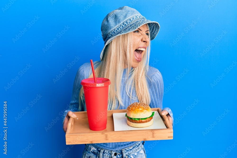 Young blonde woman eating a tasty classic burger and soda angry and mad screaming frustrated and furious, shouting with anger. rage and aggressive concept. Young blonde woman eating a tasty classic burger and soda angry and mad screaming frustrated and furious, shouting with anger. rage and aggressive concept.