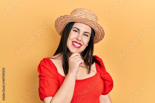 Young hispanic woman wearing summer hat looking confident at the camera with smile with crossed arms and hand raised on chin. thinking positive.