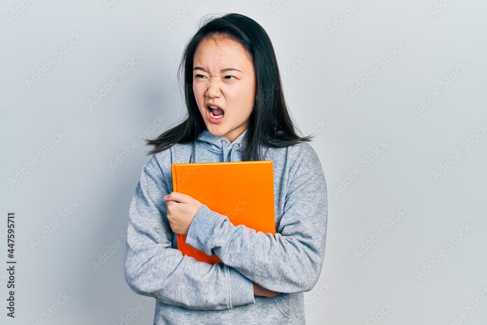 Young chinese girl holding book angry and mad screaming frustrated and furious, shouting with anger. rage and aggressive concept. Young chinese girl holding book angry and mad screaming frustrated and furious, shouting with anger. rage and aggressive concept.