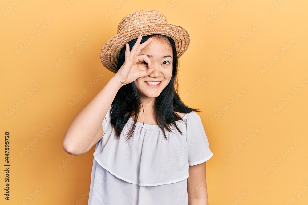 Young chinese girl wearing summer hat smiling happy doing ok sign with hand on eye looking through fingers Young chinese girl wearing summer hat smiling happy doing ok sign with hand on eye looking through fingers