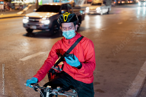 Fototapeta Naklejka Na Ścianę i Meble -  Stock photo of a food deliveryman in red uniform carrying a food delivery box to deliver for customer for order during COVID-19 pandemic and  lockdown in the city at night time in Thailand.