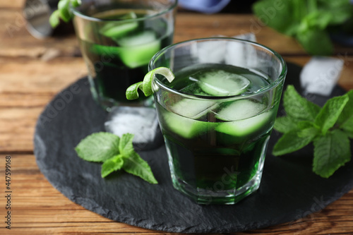 Delicious mint liqueur with ice cubes and green leaves on wooden table, closeup